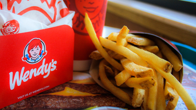 A closeup of Wendy's french fries on a tray with a disposable Wendy's burger holder and a drink on the side