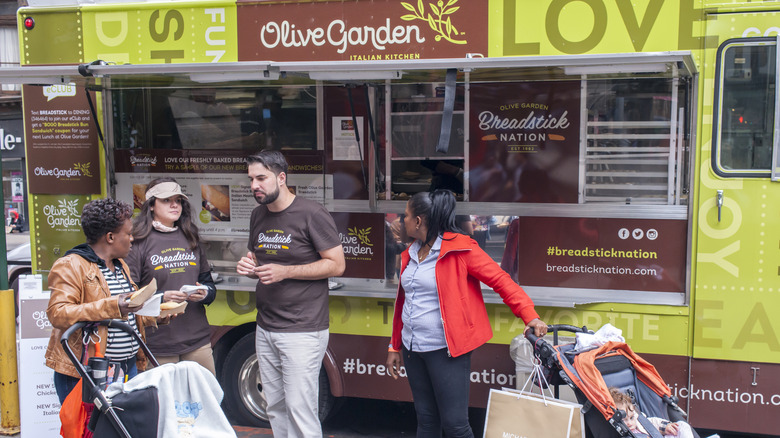 A parked Olive Garden food truck behind two female customers speaking with two Olive Garden staffs wearing Breadstick Nation t-shirts