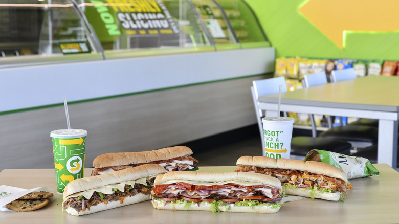Four subway sandwiches, two drinks, and cookies on a table inside a Subway store
