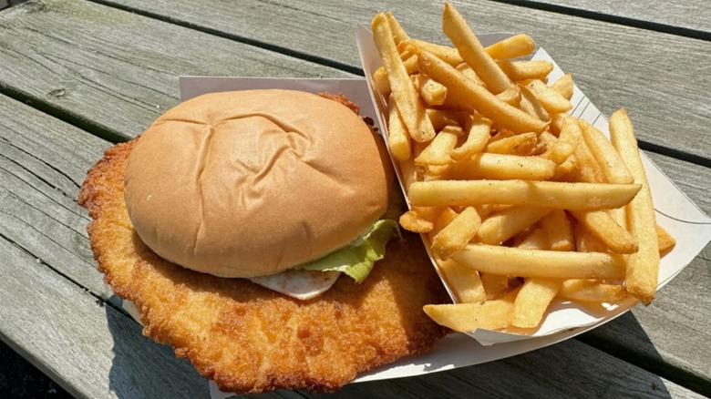 A pork tenderloin sandwich with a side of fries in a cardboard container on a wooden table