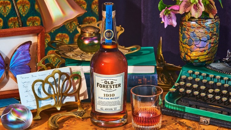 Bottle of Old Forester 1910 and glass on table with flowers, typewriter, and mounted butterfly