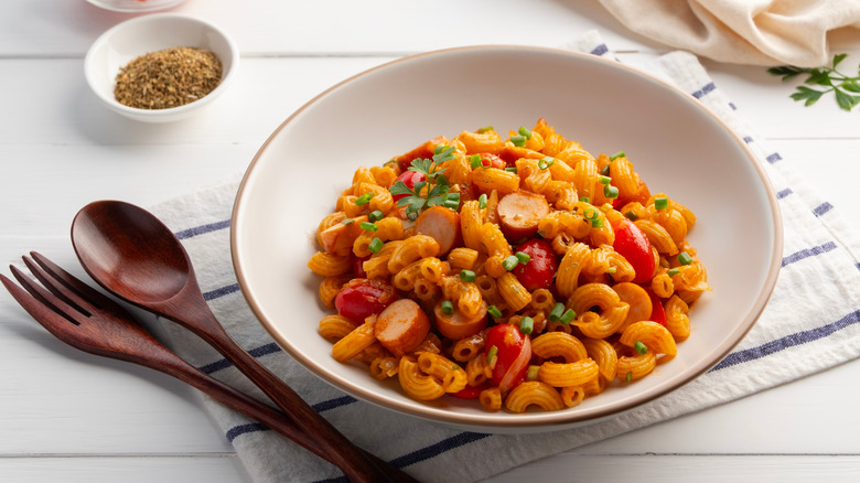 macaroni and hot dog with vegetables in a white pasta bowl with wooden cutlery on white cloth napkin