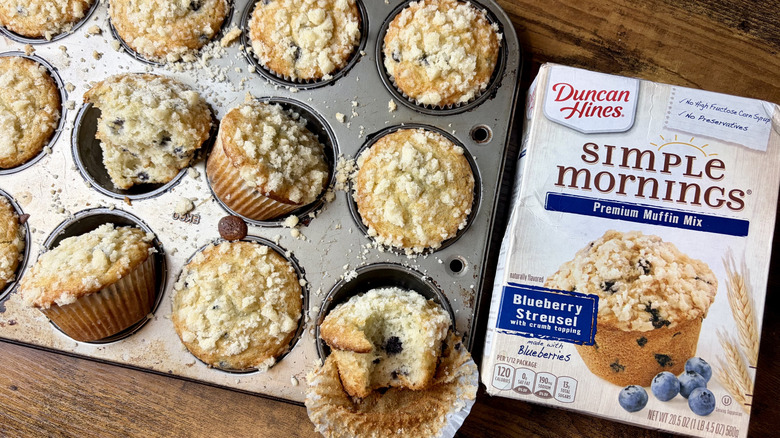 A box of Duncan Hines Simple Mornings Blueberry Stresuel Muffin Mix and a muffin pan with fresh baked blueberry muffins