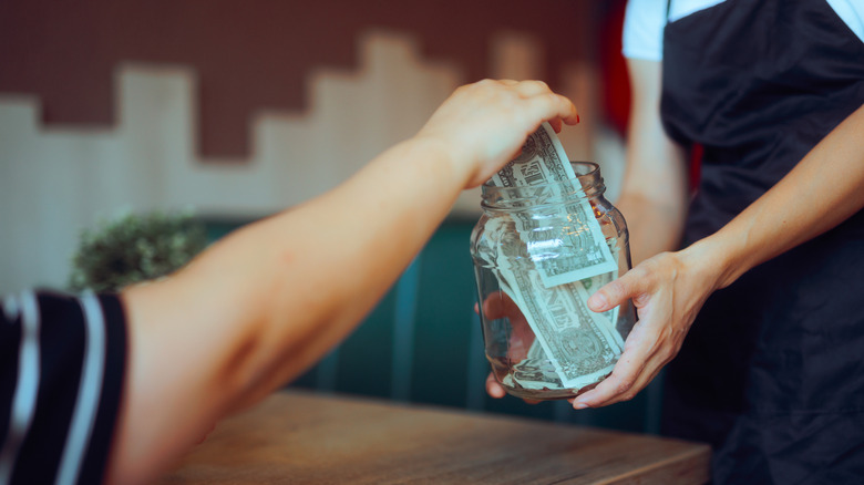 A hand of a customer putting dollars inside a tip jar held by a server