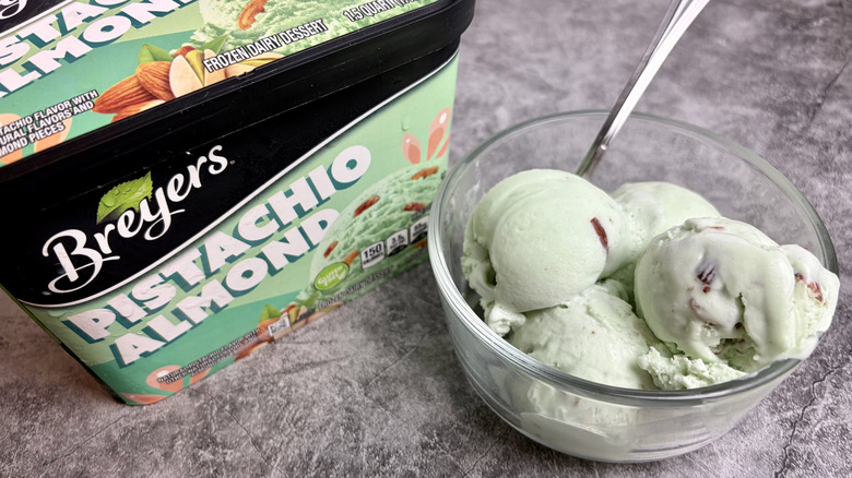A carton of Breyers Pistachio Almond ice cream next to a clear bowl with scoops of the ice cream and a spoon