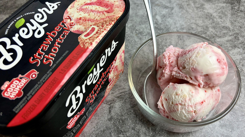 A carton of Breyers Good Humor Strawberry Shortcake ice cream next to a clear bowl with scoops of the ice cream and a spoon