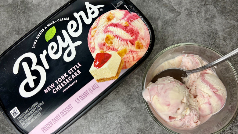 A carton of Breyers New York Style Cheesecake ice cream next to clear bowl with scoops of the ice cream and a spoon