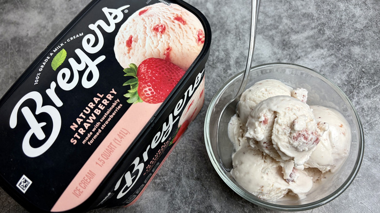 A carton of Breyers Natural Strawberry ice cream next to a clear bowl with scoops of ice cream and spoon