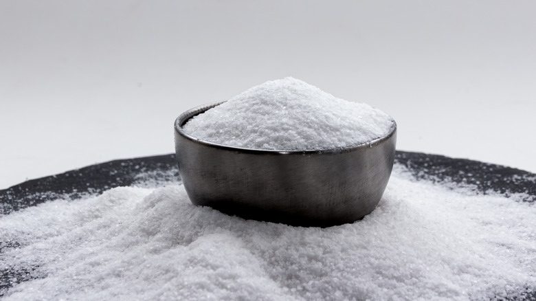 A bowl of salt surrounded by more salt on a dark surface against a white backdrop
