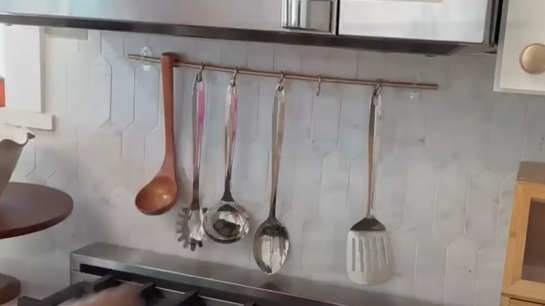 ladles, spatulas, and a pasta scooper hanging on a metal bar held up by clear adhesive hooks stuck on the backsplash above a stove