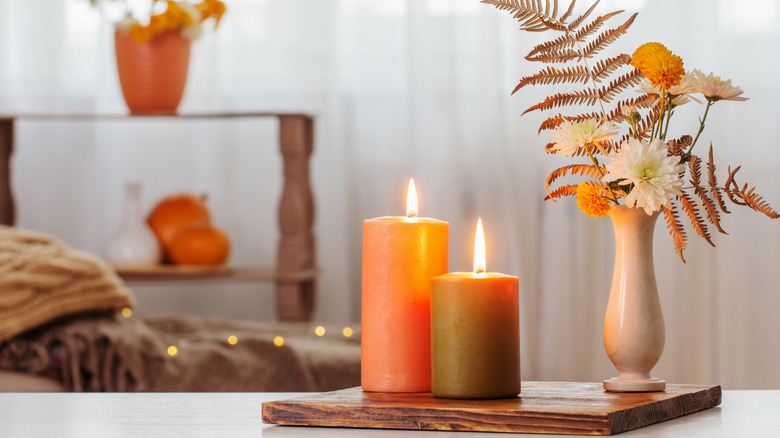 two lit candles and a tiny vase with autumn flowers stand on a wooden chopping board on a table, with knit throws and a decorative shelf with autumn-themed decorations in the background