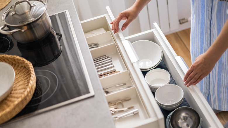 A woman's hands are visible touching two open drawers. The top drawer is filled with plastic drawer organizers with cutlery. The bottom drawer is filled with bowls.