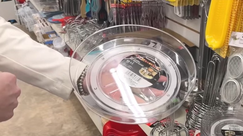 A woman's hand holding up a clear, plastic lazy Susan near an aisle inside a Dollar Tree store