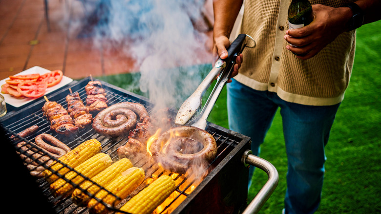 Person grilling meats and vegetables with tongs while holding bottle in the other hand.