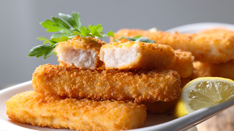 Fish sticks on a white platter wih a lemon wedge and sprig of parsley