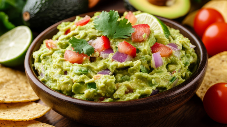close up of fresh guacamole in bowl with red onion and tomatoes with avocados in background