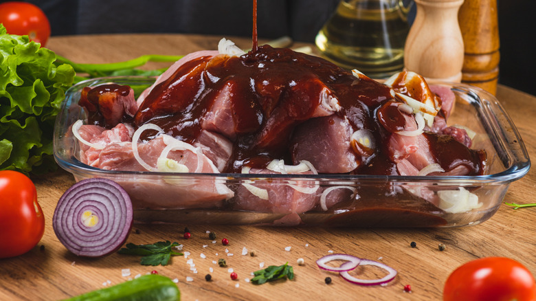Chunks of meat marinating in a glass pan surrounded by vegetables and spices on the table