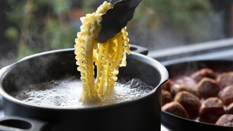 Tongs lifting pasta out of a boiling pot next to a pan of meatballs in tomato sauce