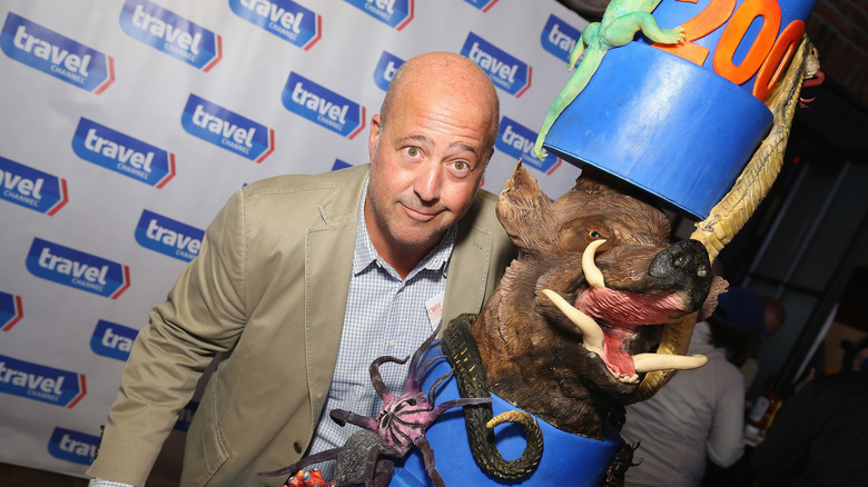 Andrew Zimmern posing next to a giant cake decorated with various wild animal sculptures celebrating "Bizarre Foods"