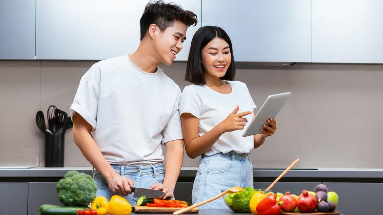 A young man and woman standing in front of two cutting boards filled with produce reading on a tablet