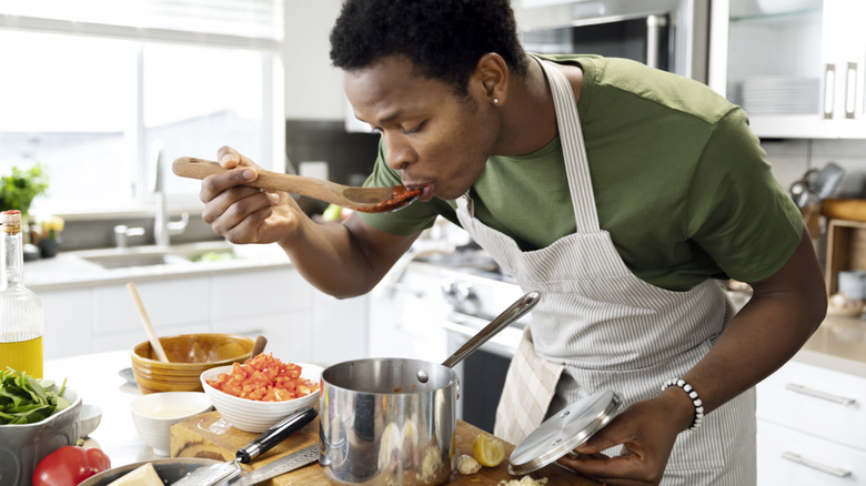 A young man bending over a pot of sauce tasting it with a wooden spoon