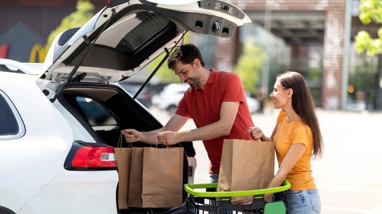 A couple loading paper bags full of groceries into their white car trunk