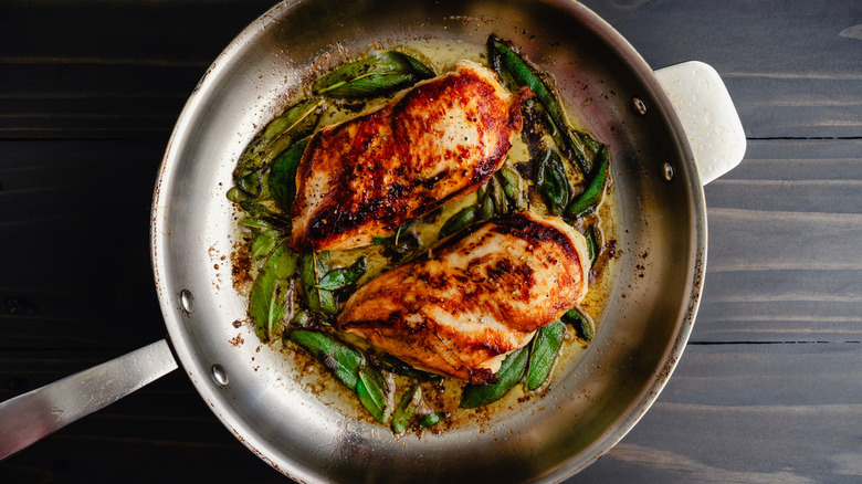 Two chicken breasts cooking in a large pan with herbs