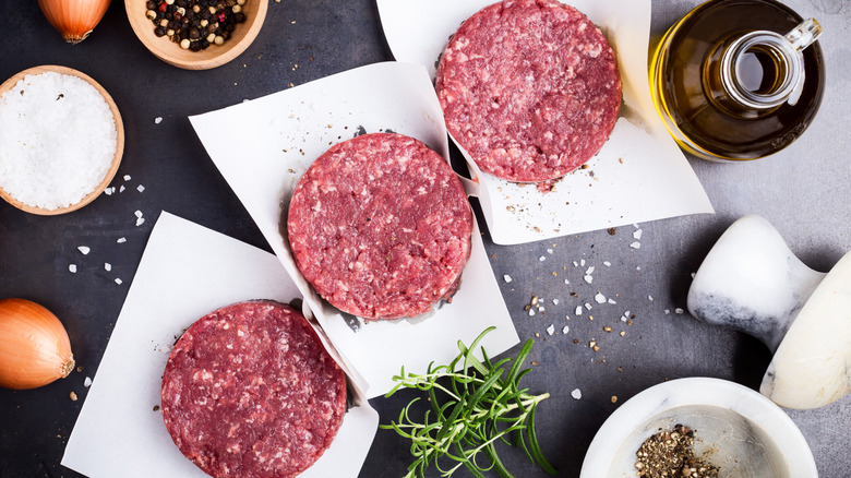 Three raw beef burger patties on paper with oil, salt, and pepper