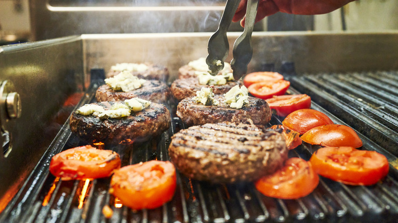 Person cooking hamburgers and sliced tomatoes on grill