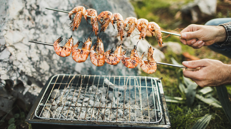 Two hands holding shrimp on metal skewers over outdoor grill