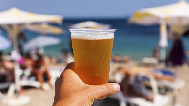 a hand holding a plastic cup of beer with the beach in the background