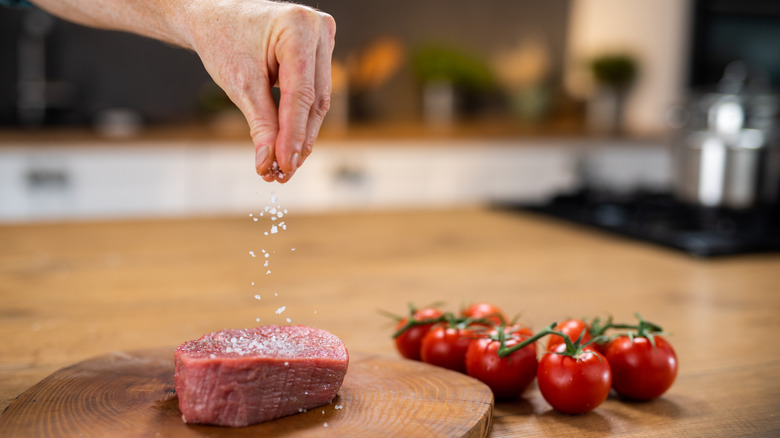 A man seasoning raw steak with salt next to tomatoes