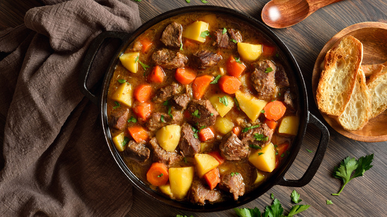 A cast iron pot of beef stew with potatoes and carrots next to a dish of slices of crusty bread and wooden serving spoon.