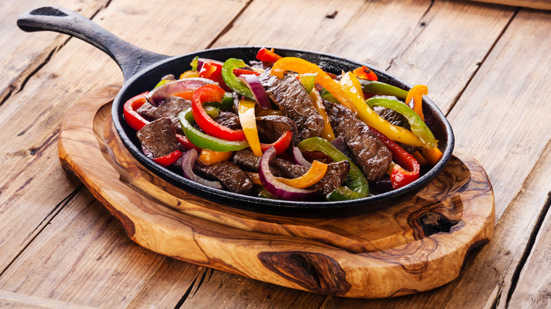 Steak fajitas with yellow, red, and green peppers in a cast-iron skillet on a wooden board and rustic wood table.