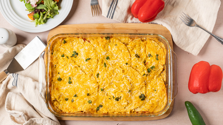 King Ranch chicken casserole in a clear, glass dish on a wooden board surrounded by a plated salad, two whole red bell and one whole jalapeño peppers, dishcloths and utensils.