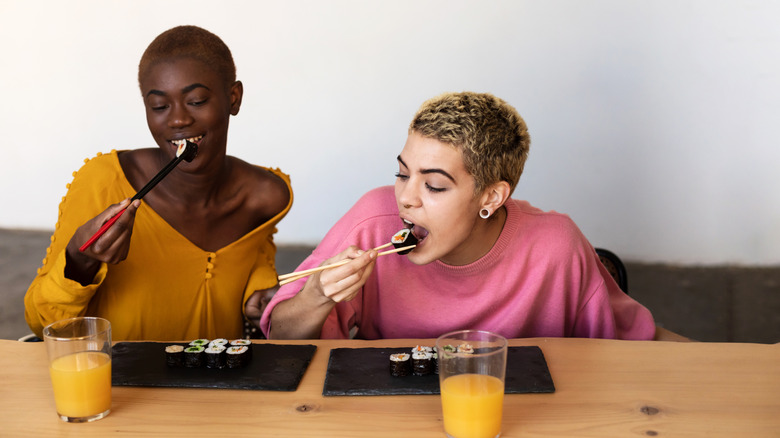 Two smiling young women eating sushi with chopsticks from stone trays with glasses of orange juice