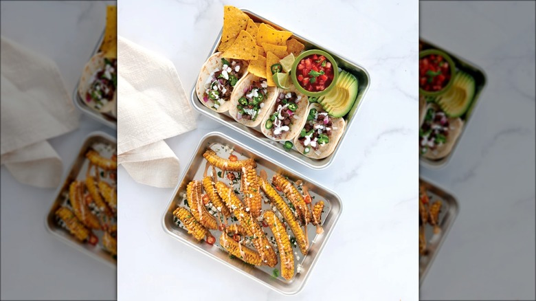 Two aluminum sheet pans loaded with street corn, tacos, and nachos.