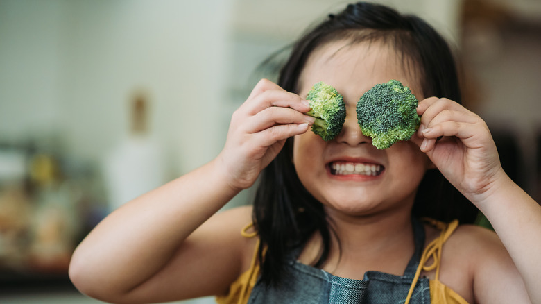 A girl covering her eyes with broccoli florets while smiling