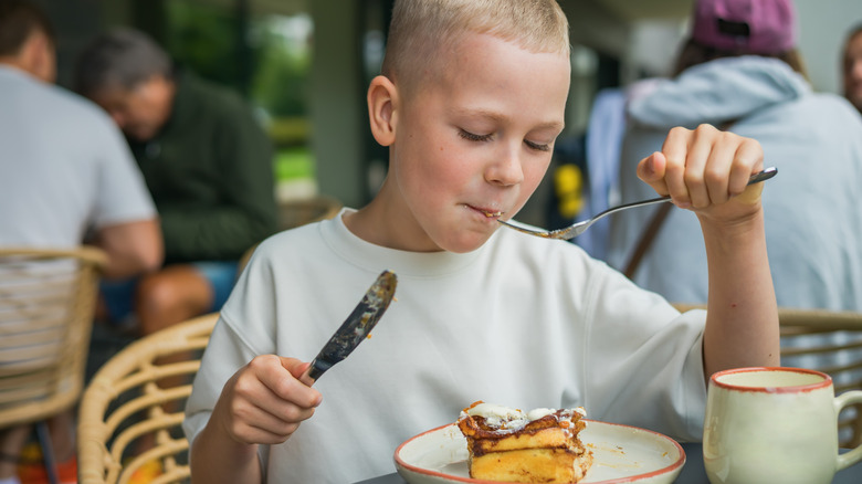 A boy eating a pastry while holding a fork and knife
