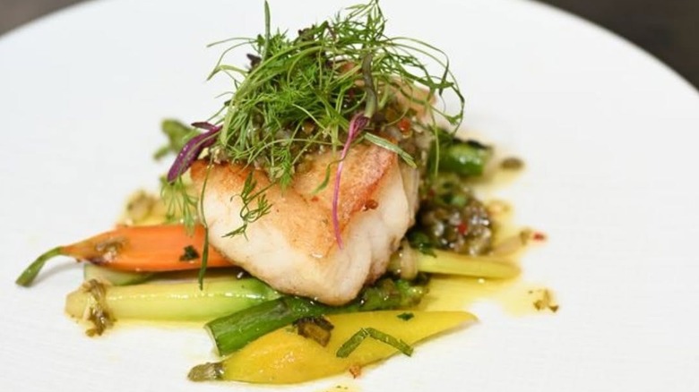 Seared white fish topped with greens on a  bed of vegetables on a white plate