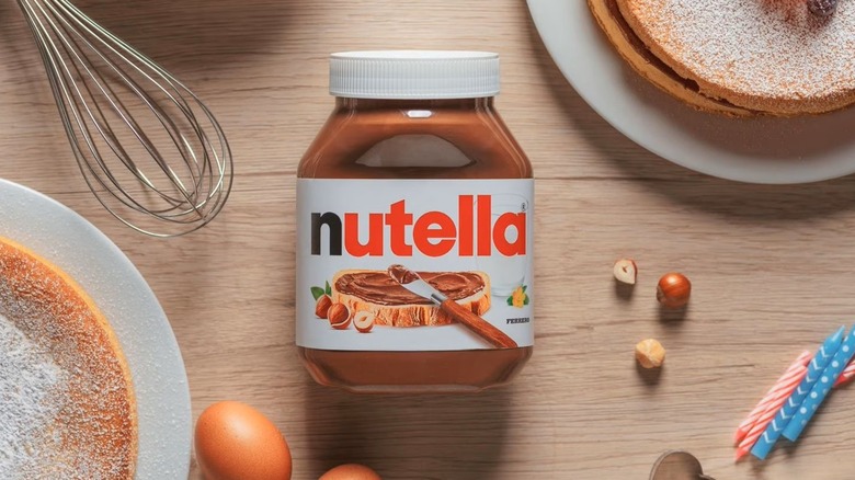 Jar of Nutella on table surrounded by crepes and other items.