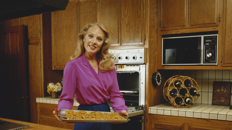1980s-inspired picture of a woman smiling at camera and holding baking sheet with food in front of open oven