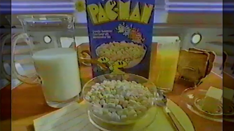 Screenshot from '80s commercial with Pac-Man cereal box in front of bowl of marshmallow cereal with glass of orange juice and glass jug of milk