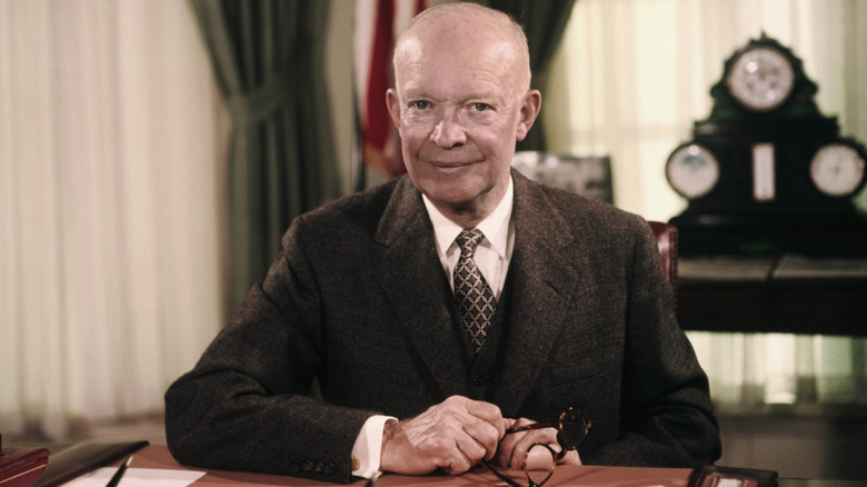 Color photo of Dwight D. Eisenhower seated at a desk in an office, wearing a suit and holding glasses