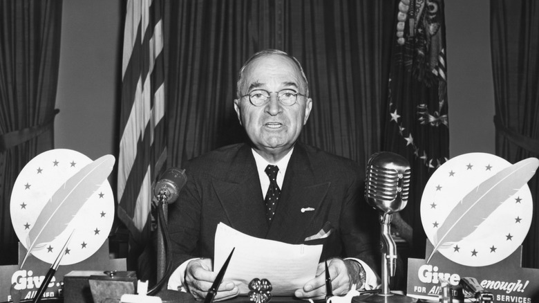 Black-and-white photo of Harry S. Truman speaking at a desk with microphones and papers