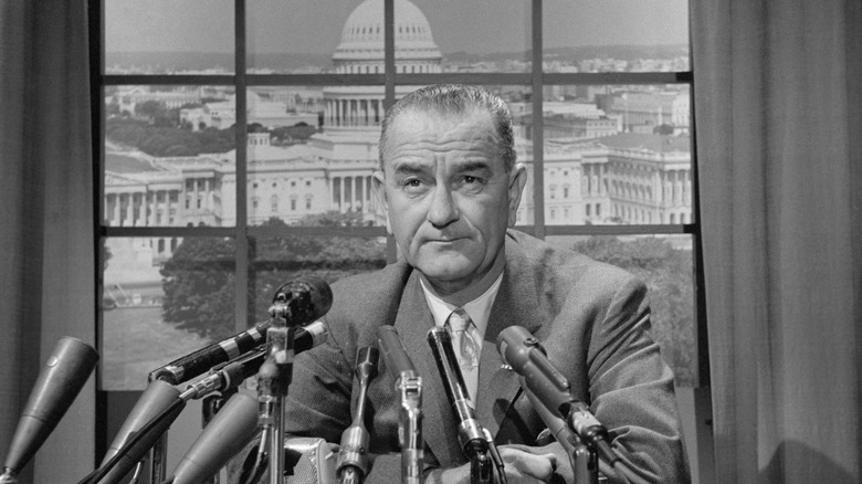 Black-and-white photo of Lyndon B. Johnson seated at a desk with multiple microphones, with the U.S. Capitol visible through a window behind him