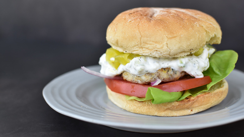 A chicken burger with lettuce, tomatoes, tzatziki sauce, red onions, and pickled banana peppers on a bun on a white plate