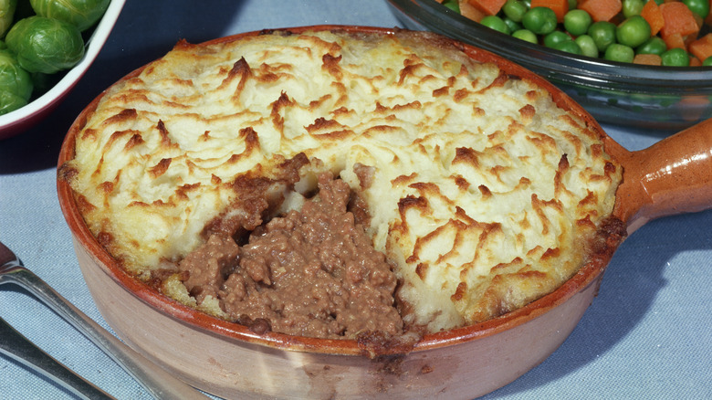 Close-up of a shepherd's pie in a ceramic pan with a bowl of peas and carrots in the background