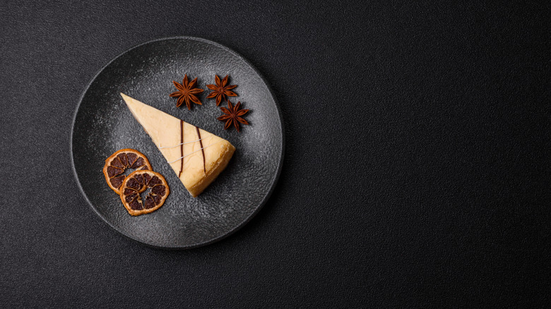 Slice of cheesecake on a black plate surrounded by dried orange slices and spices