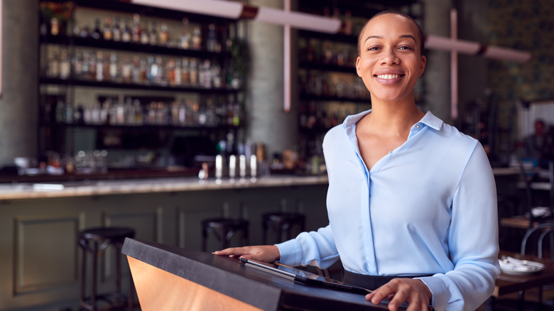 Woman standing behind restaurant check-in desk with alcohol bar in background